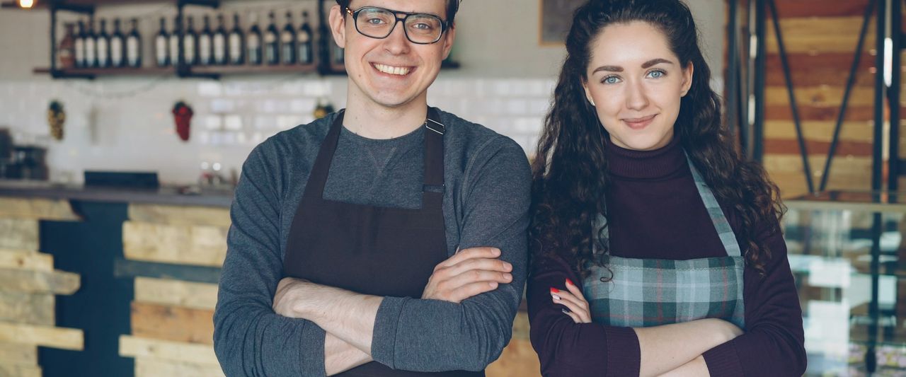 Two young cafe owners standing together inside their coffee shop, smiling