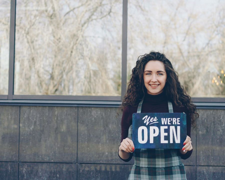 A smiling cafe worker holding a 'yes, we are open' sign outside the shop window